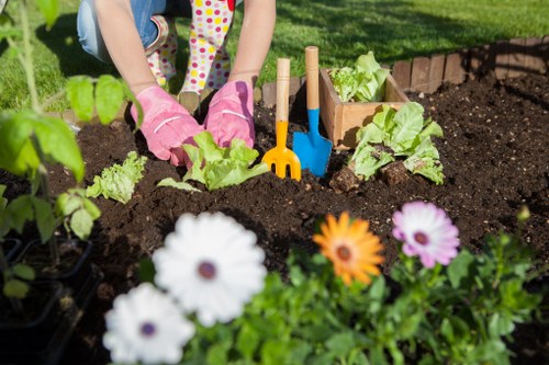 The image shows a garden scene with a child's hands, wearing pink gardening gloves, planting leafy vegetables in a prepared garden bed with dark, rich soil. Surrounding the planting area are blooming flowers, including white, purple, and orange petals, adding vibrant colour to the foreground. In the background, there is a well-maintained grassy lawn and a small border of bricks defining the garden's edge. Gardening tools such as a small blue trowel and a yellow rake are inserted into the soil, indicating active planting activity. The scene is outdoors, well-lit by natural sunlight, suggesting a clear day suitable for gardening. This setting exemplifies typical features of residential gardens in Plaistow, with a focus on vegetable planting and flower displays, aligning with professional gardening and landscaping services offered by companies like Hedge Trimming Plaistow.