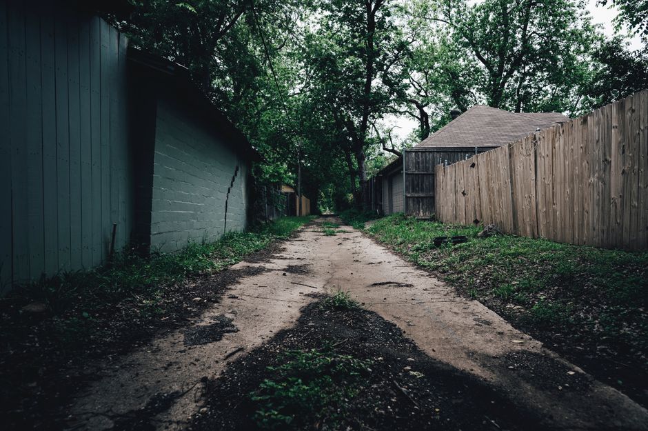 A narrow, unpaved driveway with uneven, cracked soil and patches of damp earth, bordered by tall wooden fences on both sides. To the left, a grey brick wall with visible cracks and a small cracking se
